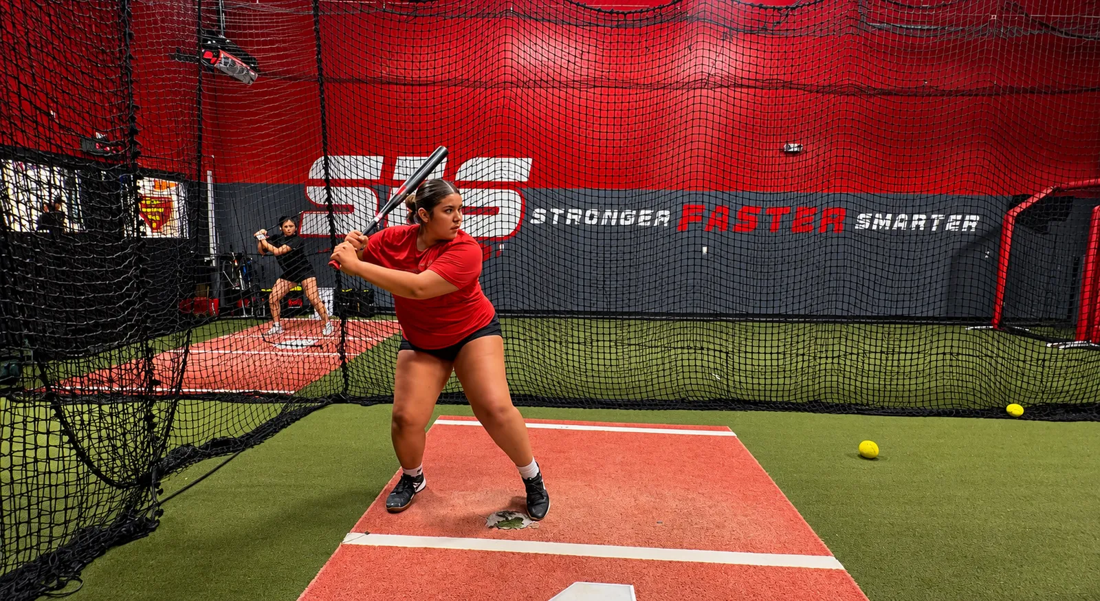 A softball hitter in her stance in the SFS hitting tunnel, with the Stronger Faster Smarter wall behind her